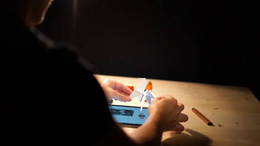 À l'atelier, des mains plient un oiseau en papier au-dessus d'une tablette posée sur un établi.